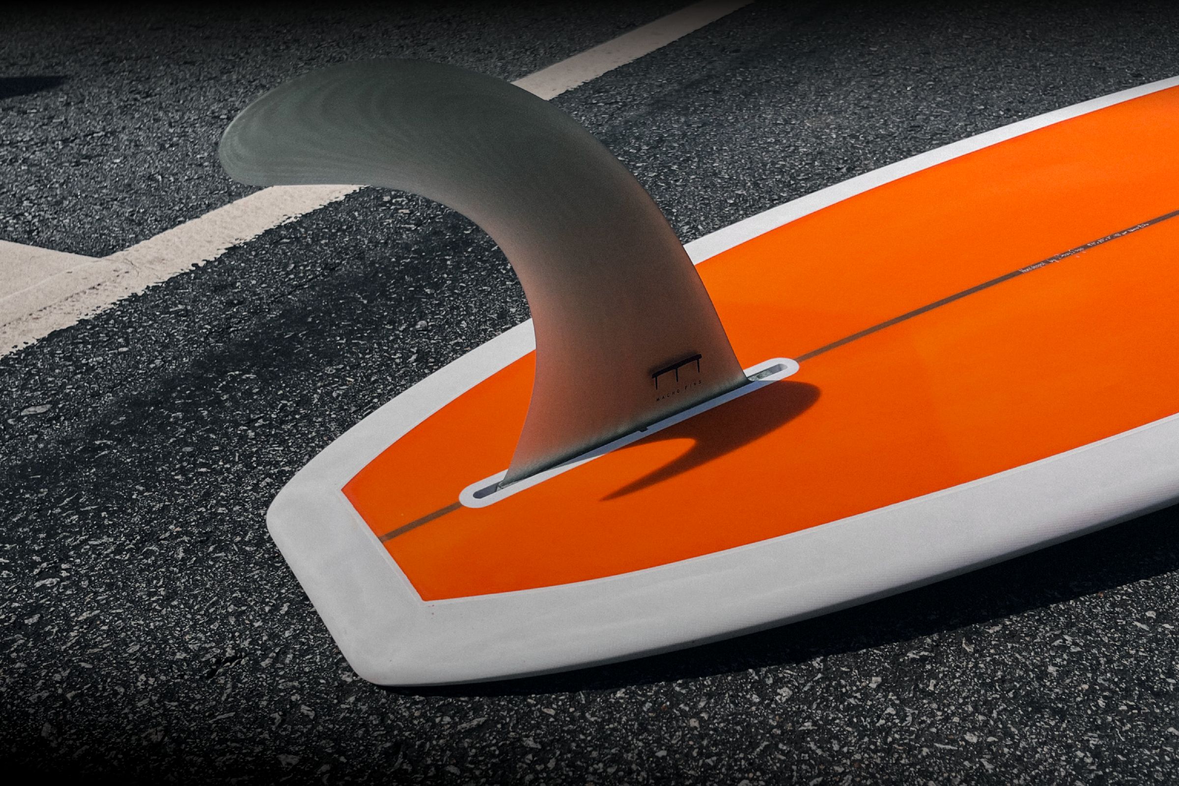 Single fin on the tail of an orange and white handmade longboard, lying on asphalt